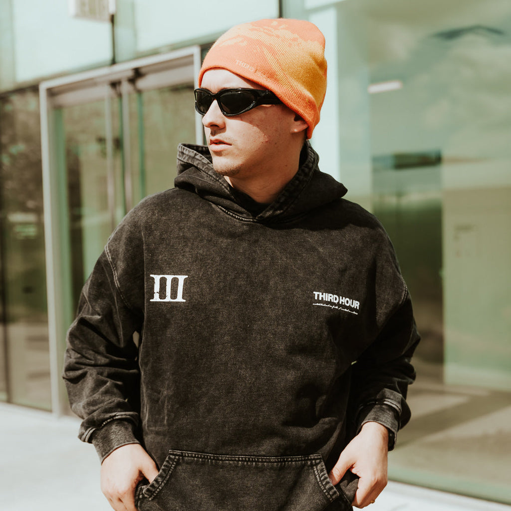 Person wearing a black hoodie with logos and an orange beanie outdoors.